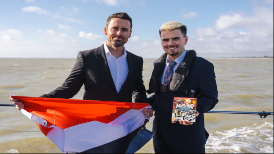Two men stand on a shore, one holds a orange, black and white flag and the other holds a copy of Metaphor ReFantazio